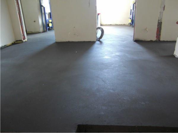 Szara, gładka wylewka podłogowa w surowym wnętrzu z białymi ścianami. Widoczne ślady po pracach remontowych i rury w tle. Surowy stan wykończenia.