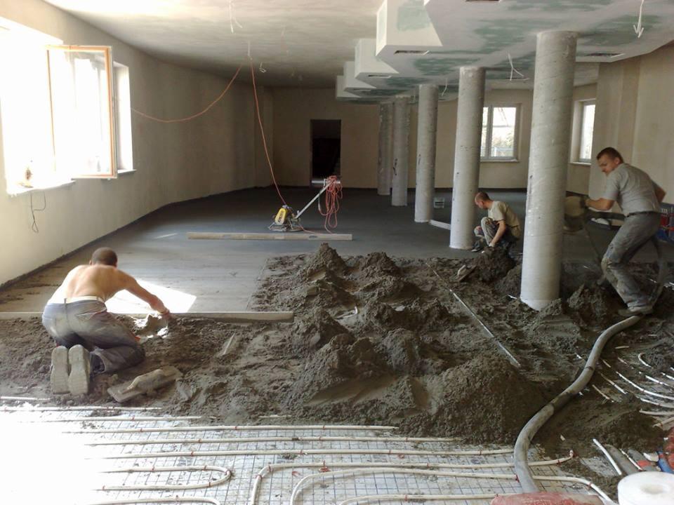 Wnętrze w trakcie prac związanych z wylewką, widoczni pracownicy, świeża wylewka oraz instalacja ogrzewania podłogowego. Jasne pomieszczenie z kolumnami.