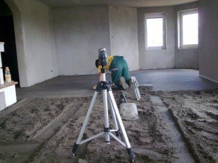 Wyrównywanie wylewki w pomieszczeniu z pomocą poziomicy laserowej na statywie i łaty, widoczny piasek i fragment gotowej podłogi.