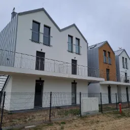 Nowoczesne, szeregowe domy z balkonami i metalową elewacją, ogrodzone siatką, z widocznymi skrzynkami gazowymi i hydrantem.