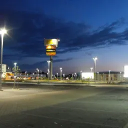 Nocny widok parkingu z oświetleniem LED, słup reklamowy Media Expert w tle. Widoczne budynki sklepowe i wieczorne niebo.