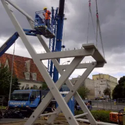 Montaż konstrukcji z betonu architektonicznego przy użyciu dźwigu. Pracownik w pomarańczowej kamizelce nadzoruje proces na wysokości. Pochmurne niebo w tle.