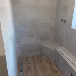 Wnętrze łazienki w trakcie wykańczania, widoczne szare płytki na ścianach, fragment okna, drewnopodobna podłoga i zamontowany odpływ liniowy przy ścianie.