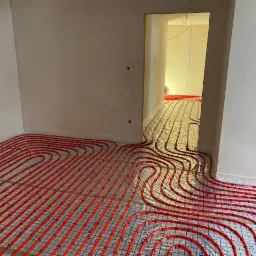 Instalacja ogrzewania podłogowego z czerwonych rurek na folii izolacyjnej w surowym wnętrzu, widok z perspektywy podłogi, z przejściem do kolejnego pomieszczenia.