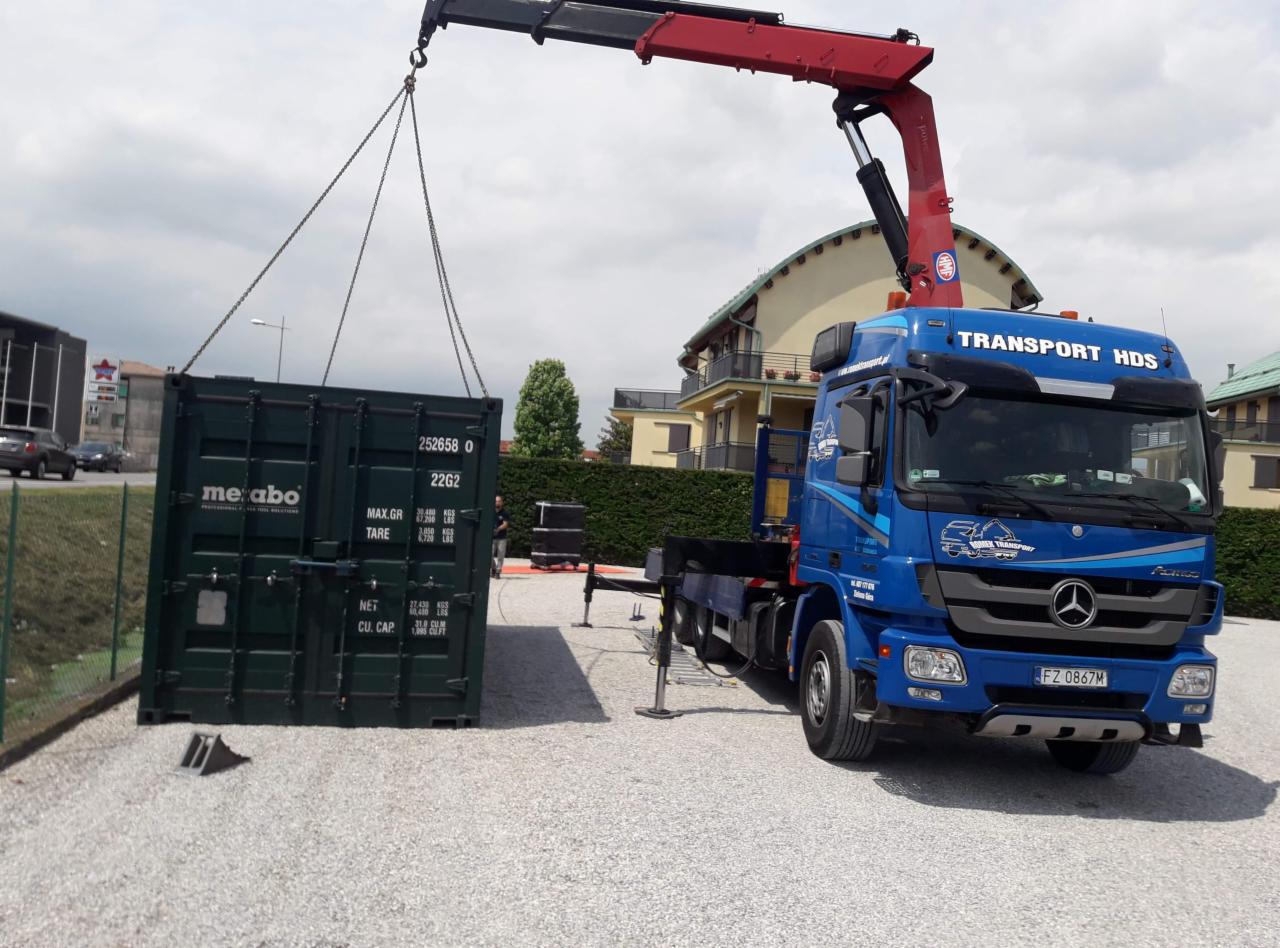 Niebieska ciężarówka z dźwigiem HDS podnosi ciemnozielony kontener Metabo na żwirowym placu, widoczne budynki w tle.