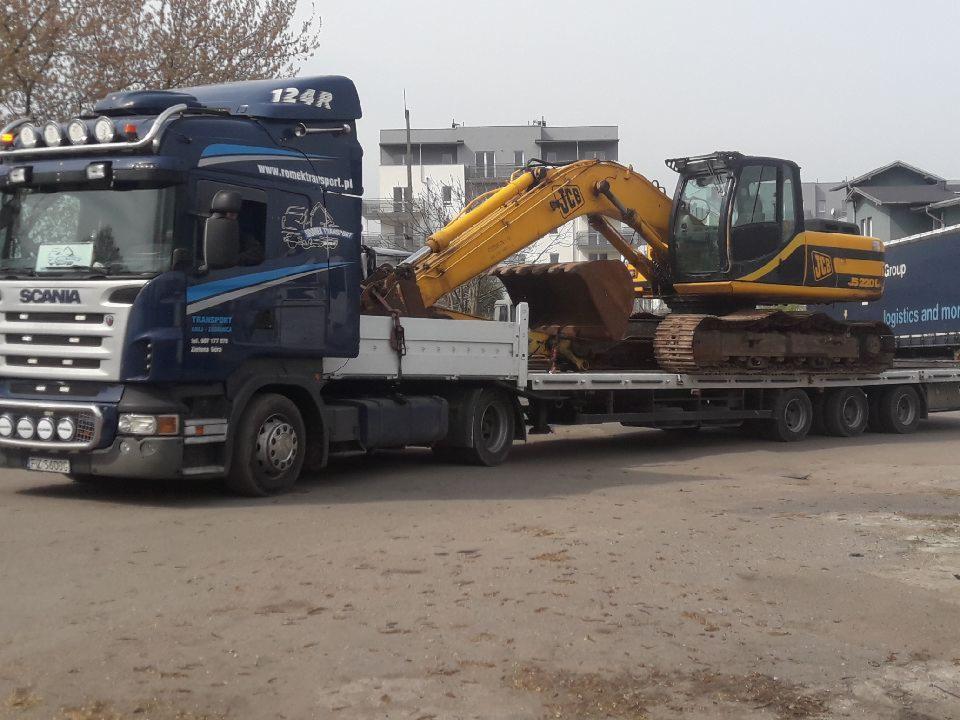 Niebieska ciężarówka Scania z naczepą niskopodwoziową przewożąca żółtą koparkę gąsienicową JCB 220L na tle budynków mieszkalnych.