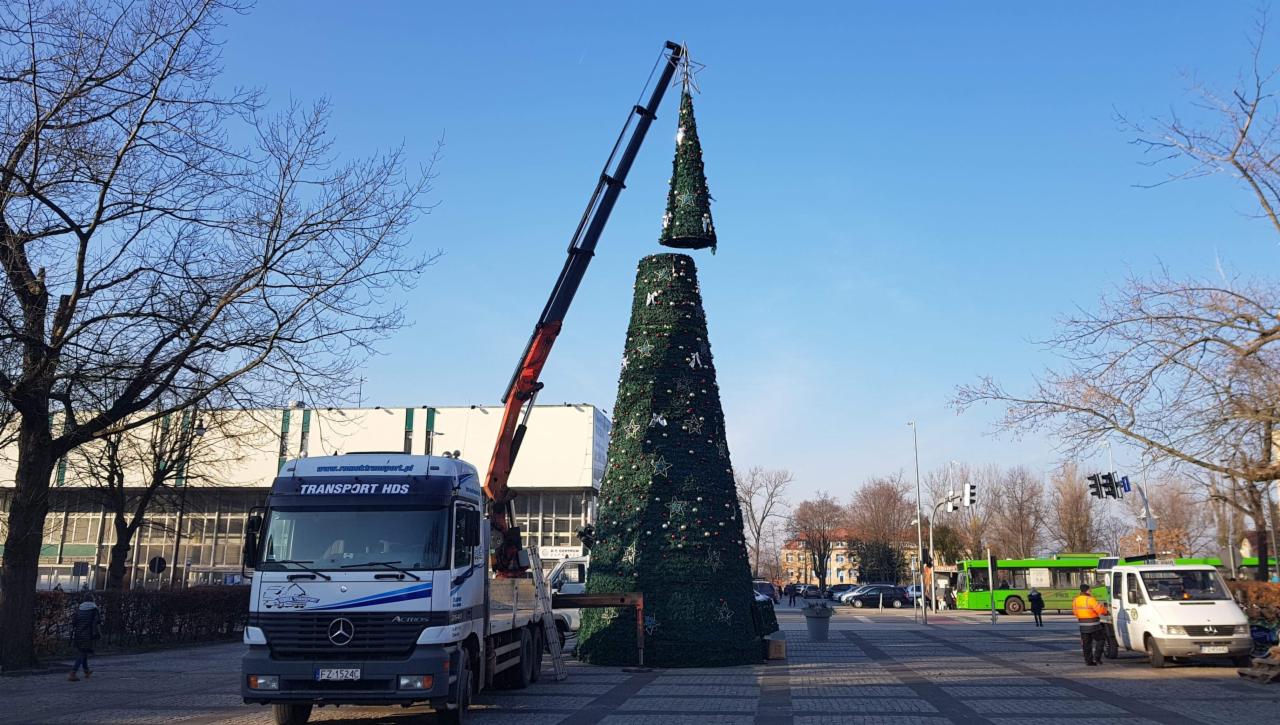 Montaż górnej części sztucznej choinki miejskiej za pomocą dźwigu HDS na placu, w tle budynek i autobus miejski.