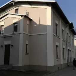 Odnowiona elewacja budynku z jasnymi, kremowymi ścianami i białymi detalami architektonicznymi, widoczne okna z ciemnymi ramami i wejście z oświetleniem, na pierwszym planie chodnik i przechodząca...