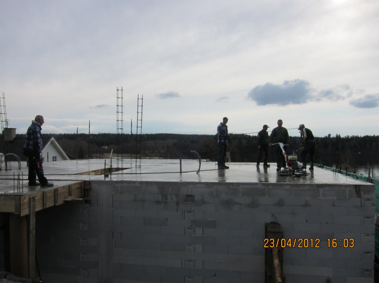 Wyrównywanie wylewki na stropie budynku w trakcie budowy. Widoczni pracownicy i maszyna do wygładzania betonu. Zbrojenie wystaje pionowo. Data w prawym dolnym rogu.