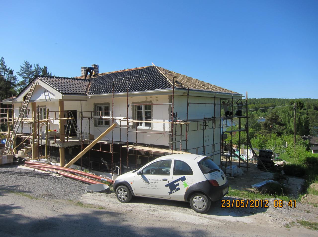 Dom w trakcie remontu: ocieplanie ścian styropianem, częściowo wymienione pokrycie dachu, rusztowania, pracownicy na dachu i rusztowaniu, firmowy samochód na pierwszym planie.