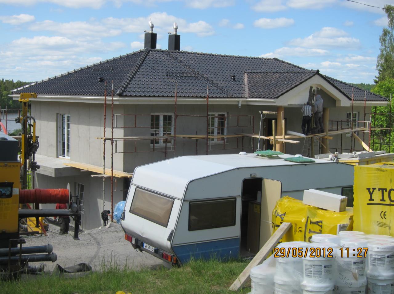 Budowa domu jednorodzinnego w trakcie tynkowania, widoczne rusztowania, przyczepa kempingowa i materiały budowlane na pierwszym planie, pochmurne niebo.