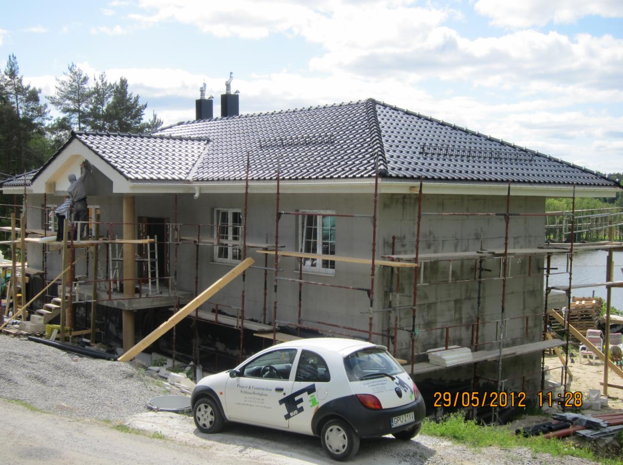 Budowa domu jednorodzinnego w trakcie prac wykończeniowych: ocieplenie, rusztowania, czarna dachówka. Na pierwszym planie auto firmy budowlanej.