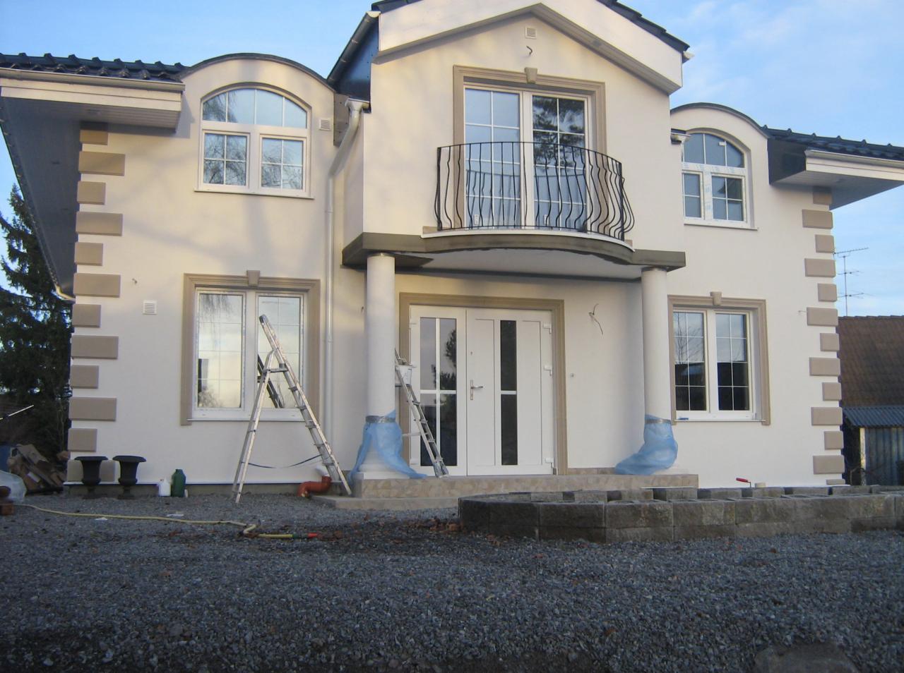 Dwukondygnacyjny dom z balkonem, w trakcie prac wykończeniowych elewacji. Widoczne drabiny, zabezpieczenia i elementy dekoracyjne. Jasna fasada i szara kostka brukowa.