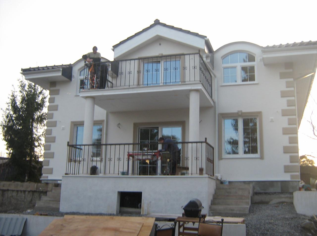 Dwupiętrowy dom z balkonami, gdzie robotnicy montują balustrady. Jeden na górnym balkonie, drugi na dolnym. Widoczne narzędzia pracy i elementy wykończeniowe.