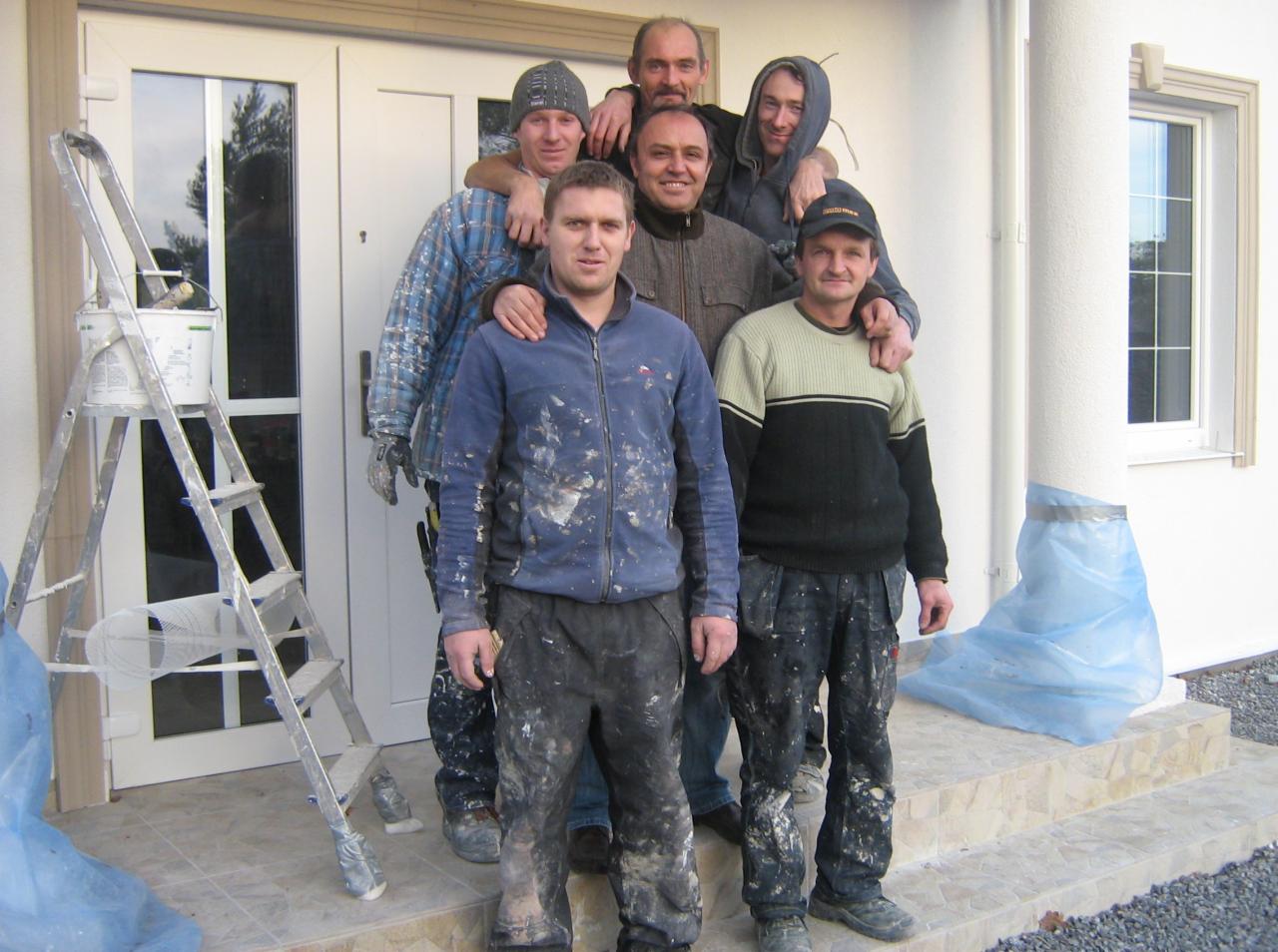 Grupa sześciu uśmiechniętych pracowników budowlanych w ubraniach roboczych, stojących przed wejściem do budynku z drabiną i wiadrem farby. Kolumna zabezpieczona folią.