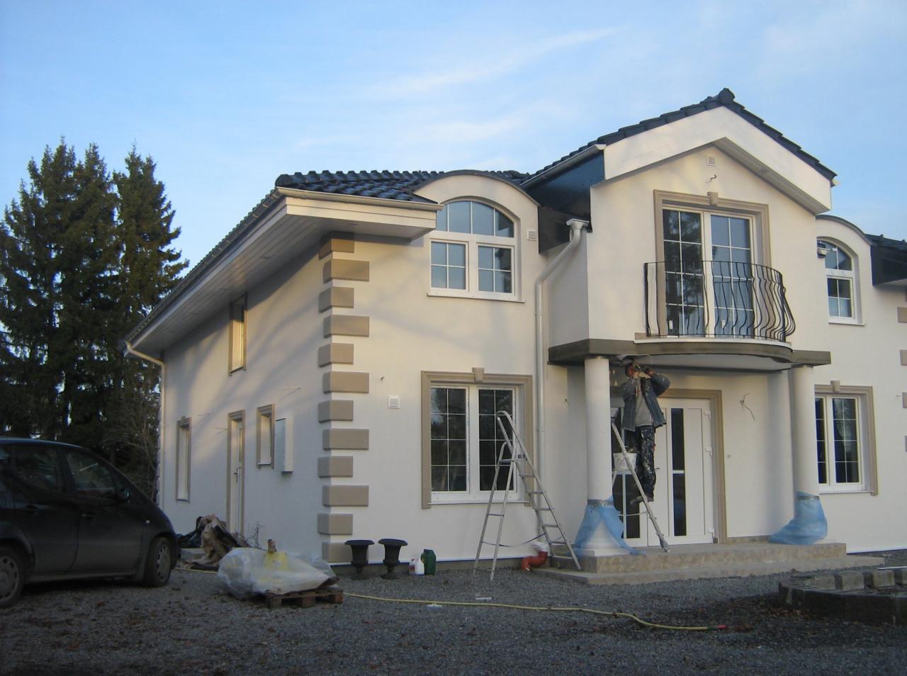 Dwupiętrowy dom z białymi ścianami i szarymi detalami. Malarz na drabinie odświeża elewację kolumn przy wejściu. Widoczne narzędzia i materiały budowlane.