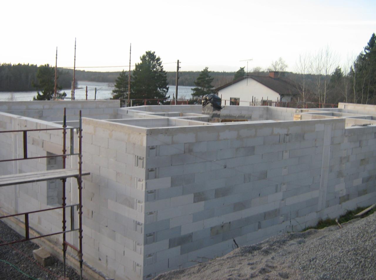 Wznoszenie ścian fundamentowych z bloczków betonowych. Widoczne zbrojenia i szalunki. W tle dom w budowie i jezioro. Prace murarskie w toku, widok z boku.