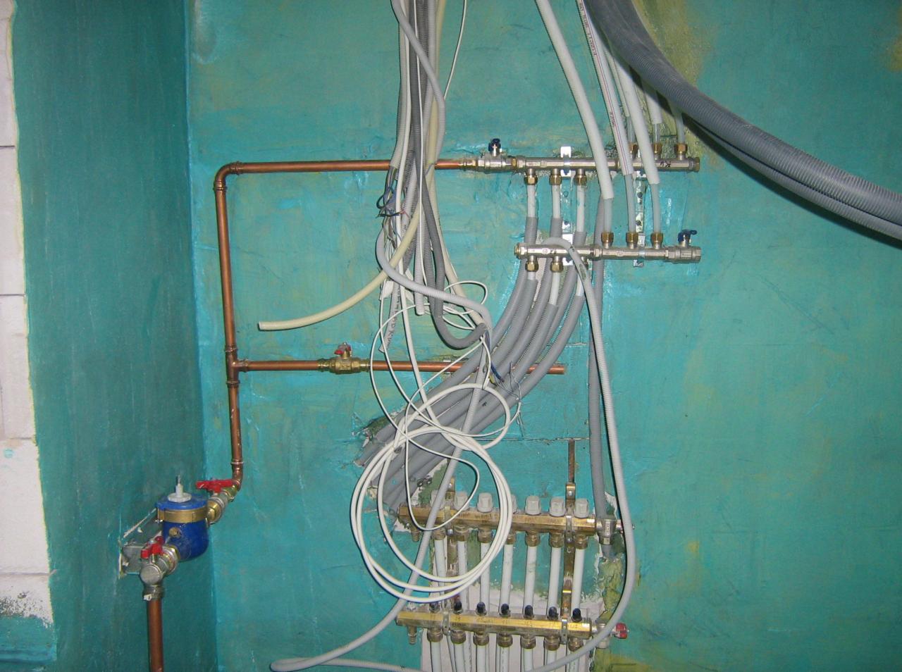 Rozdzielacz ogrzewania podłogowego z rurami na tle turkusowej ściany. Widoczne miedziane rury i liczne przewody. Instalacja w trakcie montażu.