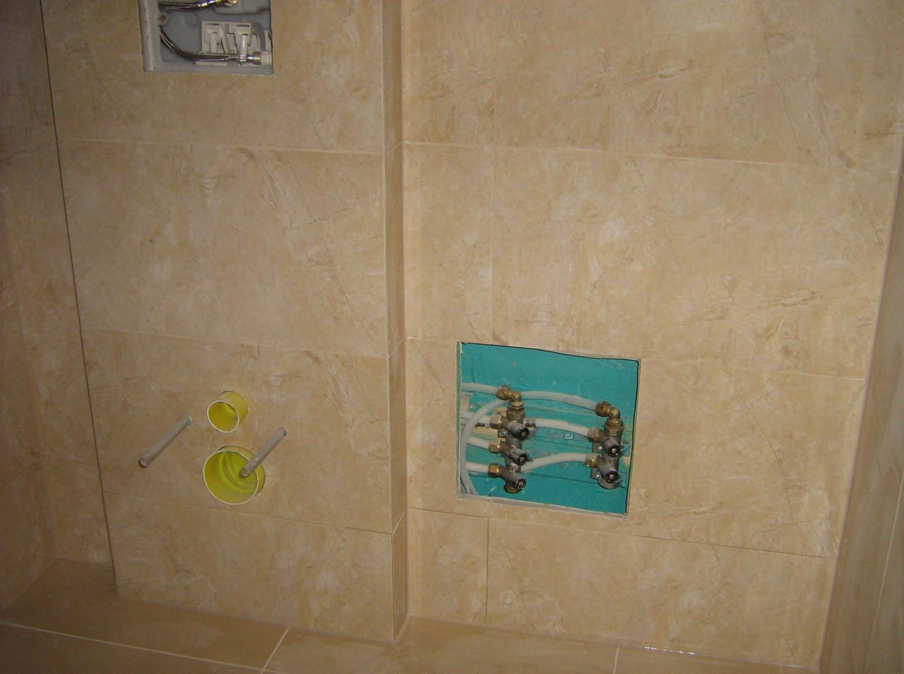 Przygotowanie instalacji wodnej w łazience z beżowymi płytkami. Widoczne rury i zawory w otworach w ścianie. Etap prac przed montażem armatury.