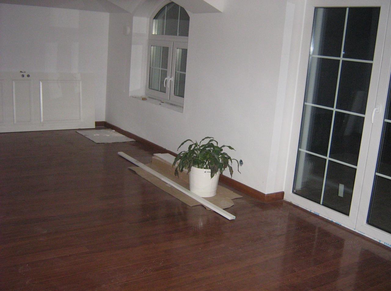 Wnętrze po remoncie: parkiet, białe ściany z oknami, listwy przypodłogowe i roślina doniczkowa. Widoczne ślady prac wykończeniowych.