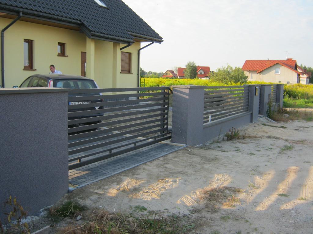 Nowoczesna, przesuwna brama posesyjna z szarymi, poziomymi panelami i słupkami, widok z zewnątrz na tle domu z żółtą elewacją i polną roślinnością.