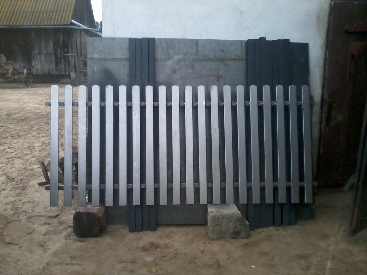 Metalowe przęsło ogrodzeniowe z pionowymi sztachetami, oparte na kamieniach, z widocznym tłem wiejskiego krajobrazu i elementami konstrukcyjnymi. Surowy wygląd.