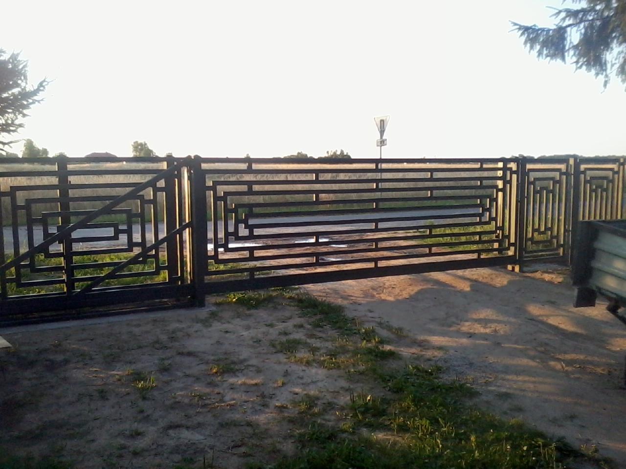 Nowoczesna, czarna brama przesuwna z geometrycznym wzorem, widziana z bliska na tle polnej drogi i zieleni. Elementy ozdobne w stylu art deco.