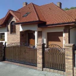 Dwukondygnacyjny dom z brązową dachówką, ceglaną fasadą, drewnianą bramą wjazdową i metalowym ogrodzeniem z dekoracyjnymi elementami roślinnymi.