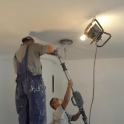 Pracownicy szlifują sufit za pomocą szlifierki teleskopowej, oświetleni lampą budowlaną. Widoczne ślady szpachli na suficie i kombinezonach.