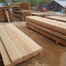 Stosy świeżo przetartego drewna budowlanego ułożone na placu, w tle budynek gospodarczy i wózek widłowy.