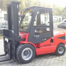 Czerwony wózek widłowy M40 marki Suchedniów na kostce brukowej, z budynkami i drzewami w tle, widziany z boku.