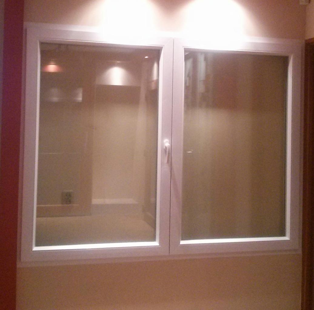 Dwuskrzydłowe okno PCV z białymi ramami, widok na wnętrze z oświetleniem punktowym. Zamknięte okno z klamką w pozycji pionowej.