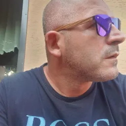 Mężczyzna z krótkimi włosami w okularach przeciwsłonecznych z fioletowymi szkłami, patrzy w prawo. Widoczny fragment okna z zasłoną.