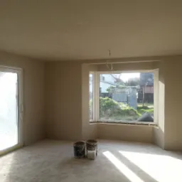 Wnętrze po tynkowaniu maszynowym: gładkie ściany w odcieniach beżu, widoczne dwa wiadra i okno z widokiem na zieleń i zabudowania.