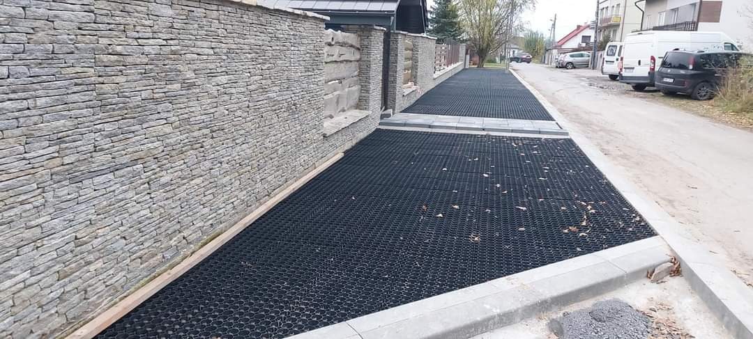 Ułożona, ażurowa nawierzchnia z tworzywa sztucznego na podjeździe, ograniczona kamiennym murem i betonowym krawężnikiem, widok z perspektywy ulicy.