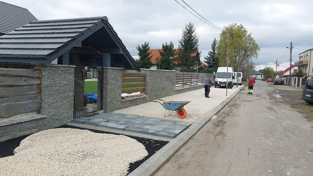 Układanie kostki brukowej w Dobryszycach, widoczny fragment podjazdu z ułożoną kostką, żwirem i pracownikami w tle. W tle zaparkowane busy.