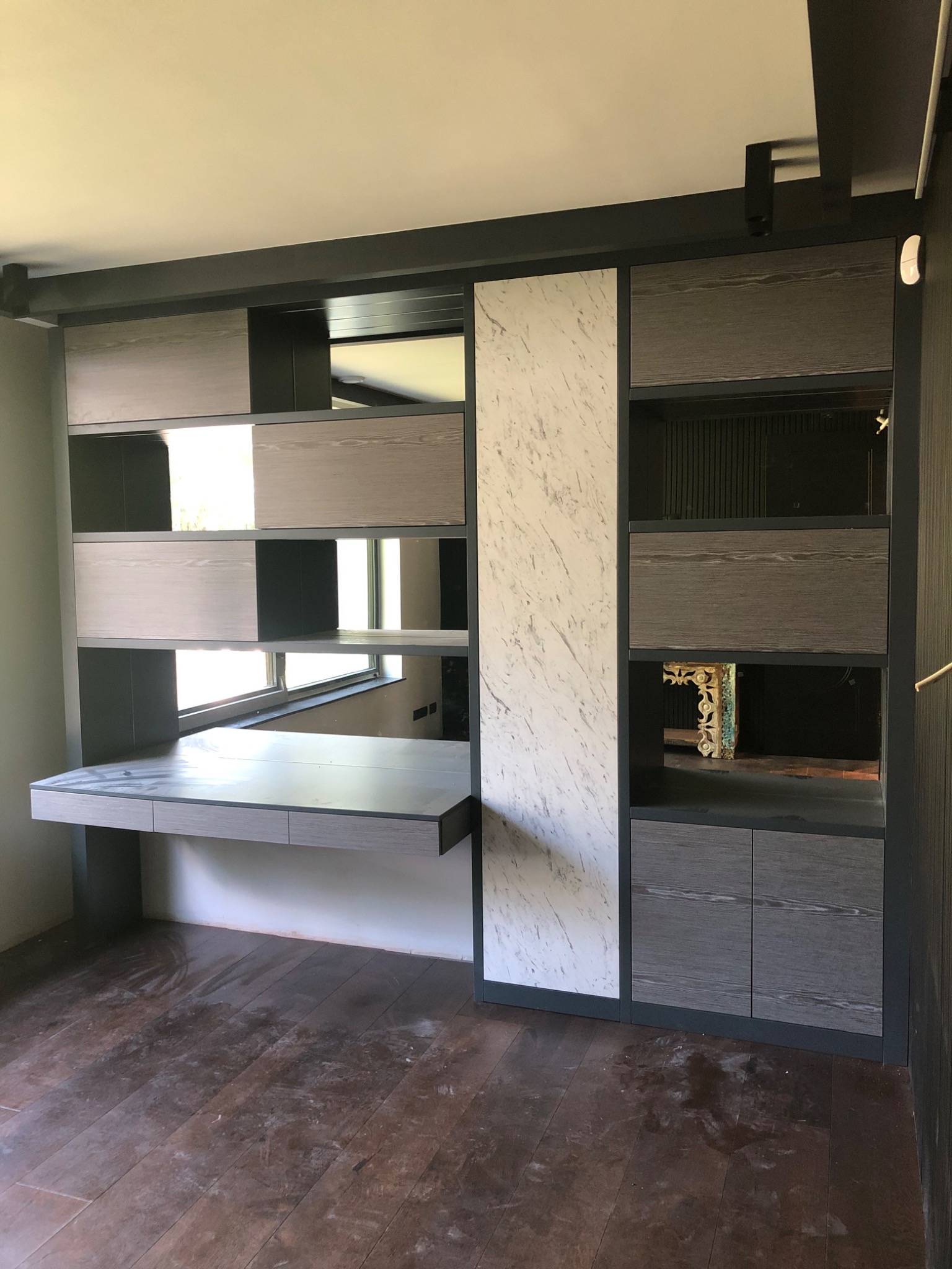 Zabudowa wnęki z szarymi szafkami z drewnopodobnym fornirem, blatem biurka i lustrzanymi wstawkami, uzupełniona panelem z imitacją marmuru.