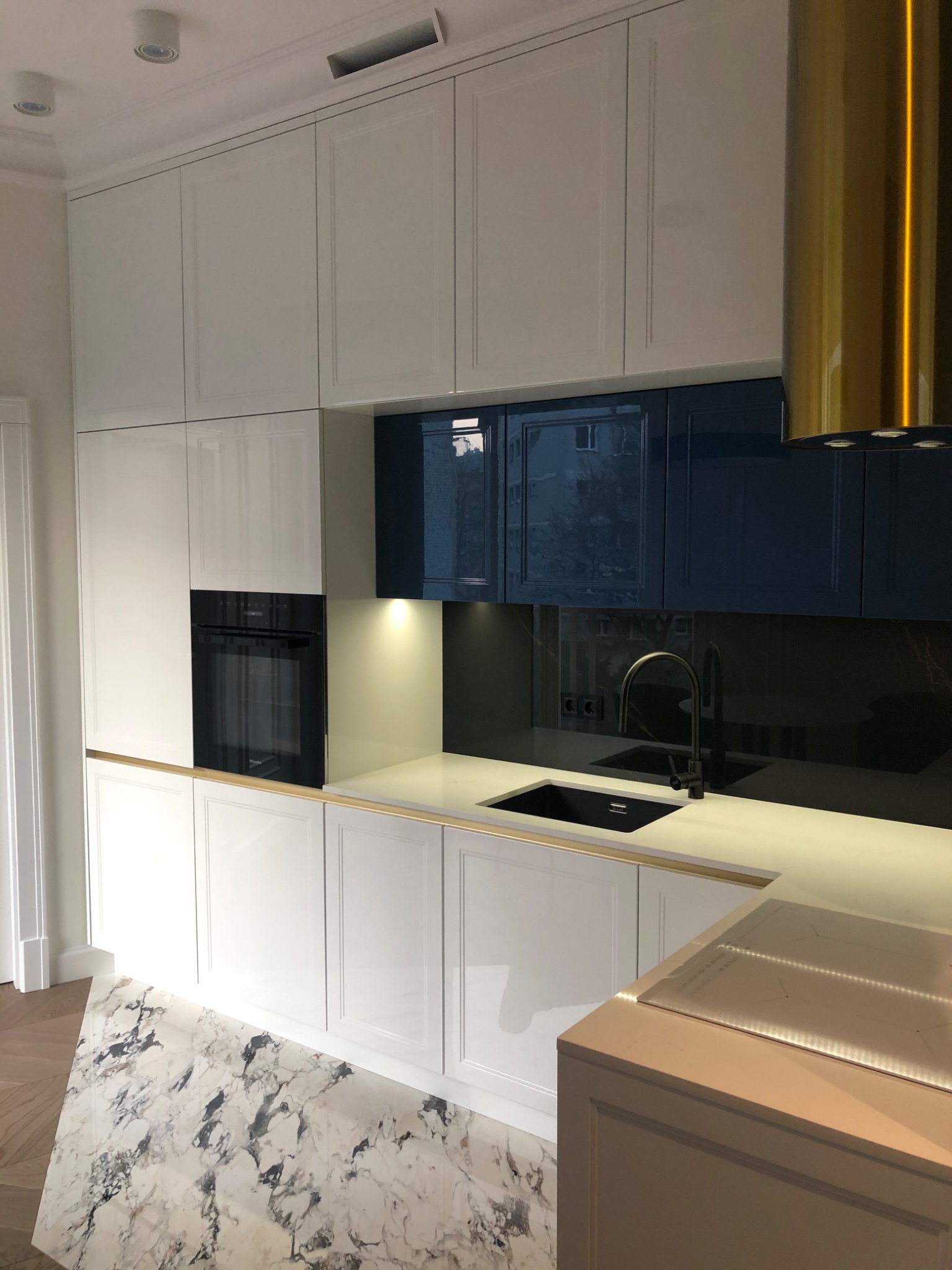 Nowoczesna kuchnia z białymi i granatowymi szafkami, złotym okapem i blatem z delikatnym złotym akcentem. Widoczny piekarnik w zabudowie i czarny zlew. Podłoga wyłożona płytkami imitującymi marmur.