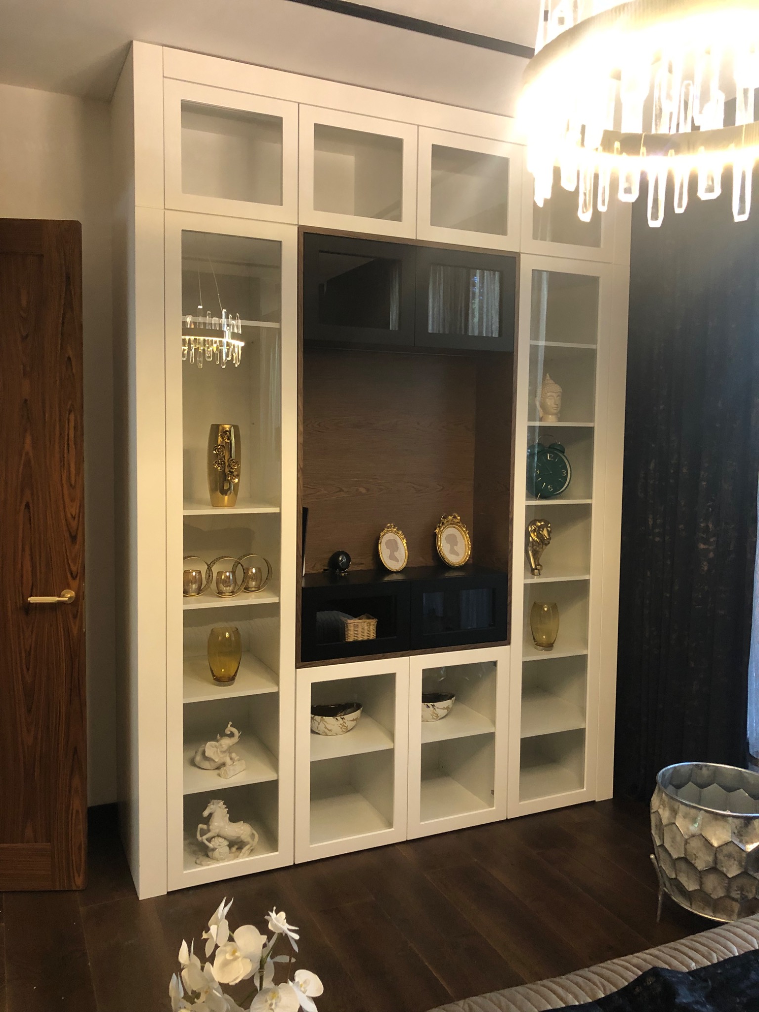 Biała, wysoka szafa z przeszklonymi drzwiczkami, wbudowana w ścianę, z dekoracjami na półkach, oświetlona wiszącym żyrandolem.