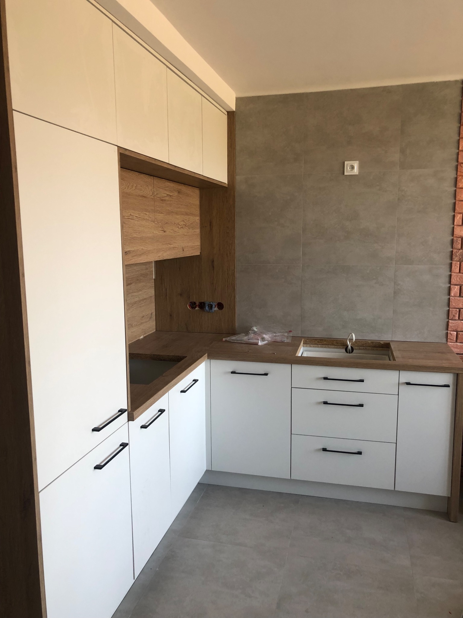 Nowoczesna kuchnia z białymi frontami szafek i drewnianymi blatami, widoczne gniazdka elektryczne na ścianie z szarych płytek, brakujące elementy wykończenia wokół zlewu.