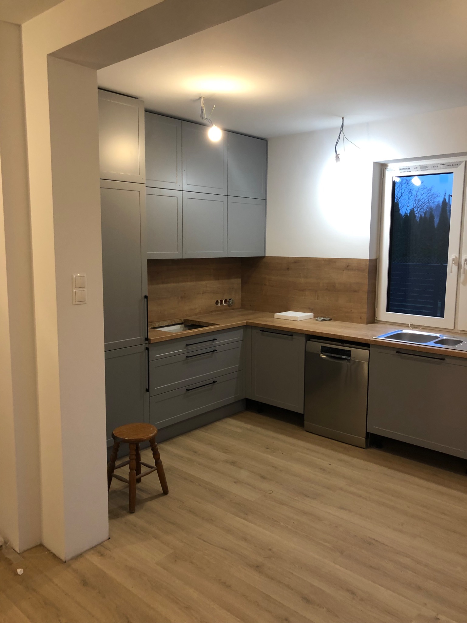 Szara kuchnia z drewnianym blatem i brązowym panelem ściennym, widoczny fragment niezakończonej instalacji oświetleniowej i drewniany stołek.