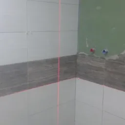 Wykończona ściana łazienki z szarymi i białymi płytkami, poziomowana laserowo. Widoczne przyłącza wodne na zielonej ścianie.