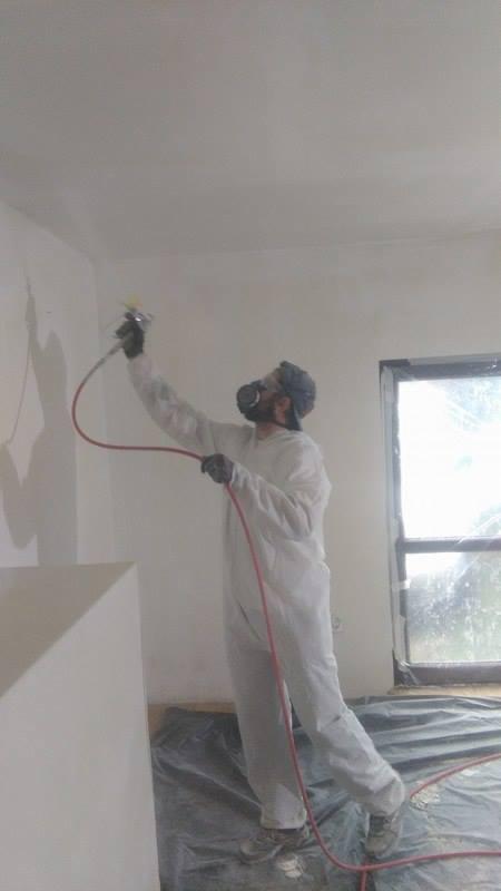 Malowanie ścian agregatem w pomieszczeniu. Pracownik w kombinezonie ochronnym i masce rozpyla farbę na ścianę. Okno w tle, podłoga zabezpieczona folią.