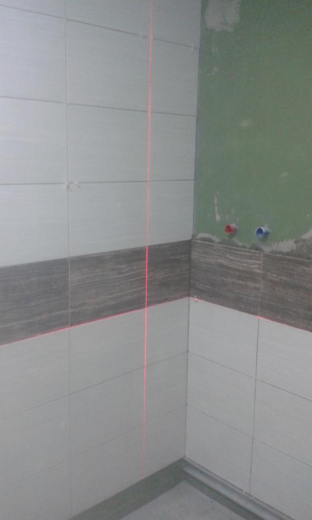 Wykończona ściana łazienki z szarymi i białymi płytkami, poziomowana laserowo. Widoczne przyłącza wodne na zielonej ścianie.