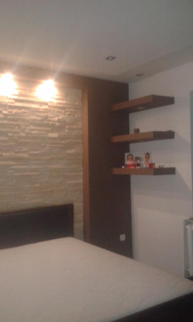 Sypialnia z łóżkiem, kamienną ścianą i półkami. Widoczne zdjęcia i dekoracje na półkach. Oświetlenie sufitowe dodaje ciepła. Aranżacja wnętrza z elementami dekoracyjnymi.