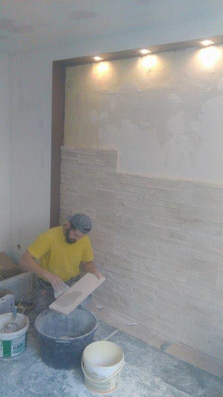 Mężczyzna w żółtej koszulce układa dekoracyjne płytki na ścianie wewnątrz pomieszczenia z oświetleniem punktowym.