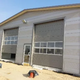 Szara hala z dwoma segmentowymi bramami garażowymi z oknami i wejściem, na piasku. Lekko uchylona brama. Ściany z poziomymi paskami. Błękitne niebo.