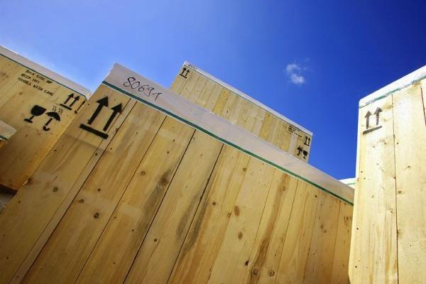 Drewniane skrzynie transportowe z oznaczeniami, ustawione pod kątem na tle błękitnego nieba. Widoczne detale drewna i czarne symbole informacyjne.