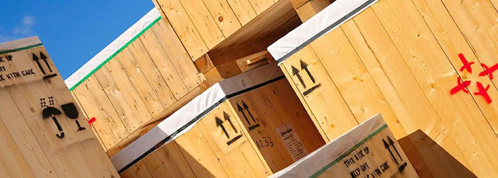 Drewniane skrzynie transportowe z oznaczeniami 'Góra' i 'Ostrożnie', ustawione pod kątem na tle błękitnego nieba. Widoczne detale: czerwone krzyże na jednej ze skrzyń.