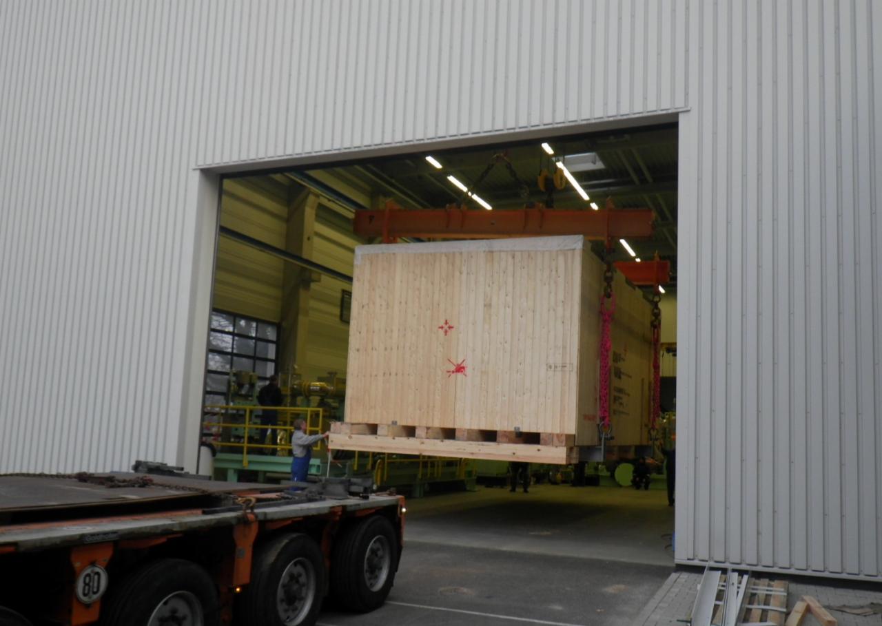 Transport skrzyniowej konstrukcji na palecie za pomocą suwnicy do wnętrza hali magazynowej, pracownik asekuruje ładunek.